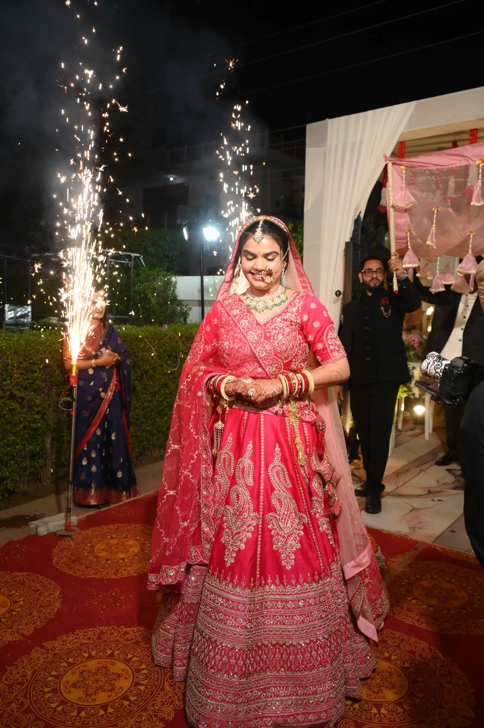 Bride in a red lehenga walking down the aisle between cold-pyro sparklers at a night wedding