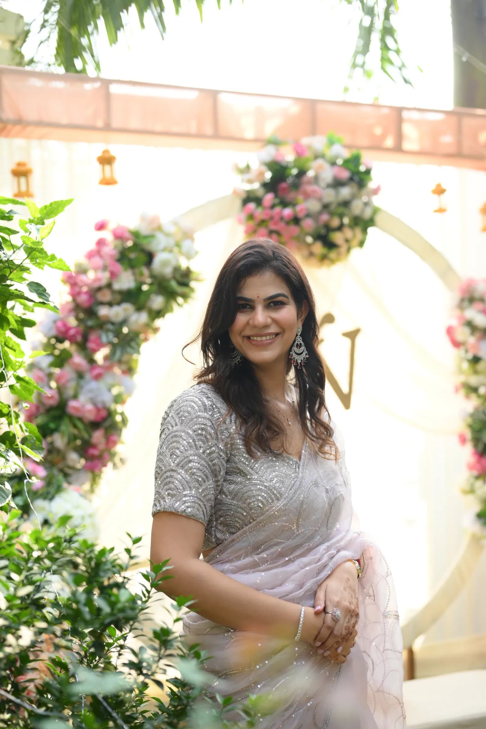 Woman in a silver saree smiling in front of a floral arch