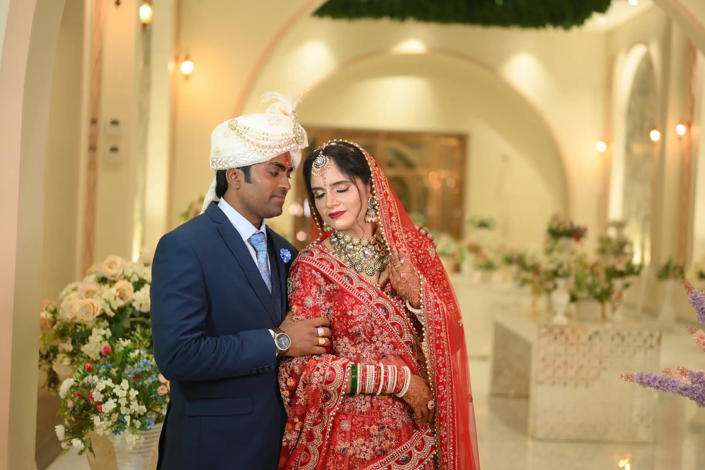 Groom in a blue suit and safa with the bride in a red lehenga in a floral-decorated hall